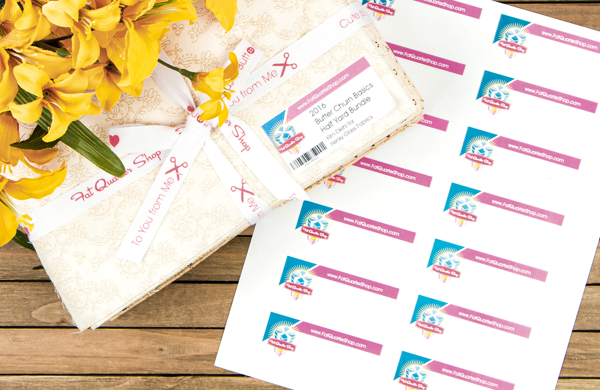 A sheet of 14 blank product labels. They have a blue corner with a logo that reads Fat Quarter Shop and a pink bar across the top that reads Fat Quarter Shop dot com. A filled in label with a barcode is stuck to a beige bundle of fabric with yellow flowers resting on top. class=
