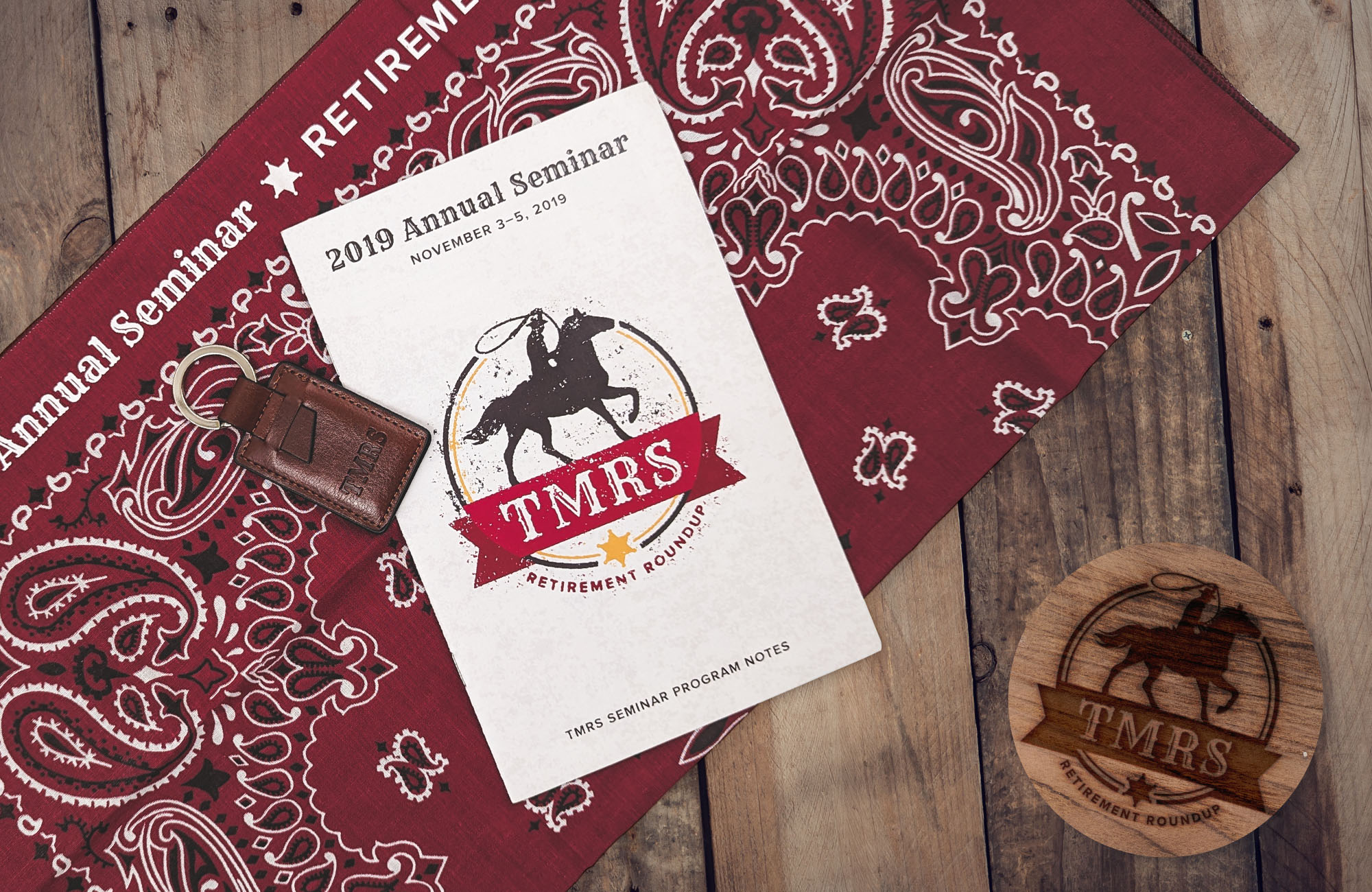 An event program for the 2019 TMRS Annual Seminar next to a leather keychain on top of a red bandana. The logo, which looks like a cowboy with a lasso, appears to be burned into  a wooden coaster in the bottom right of the photo. class=