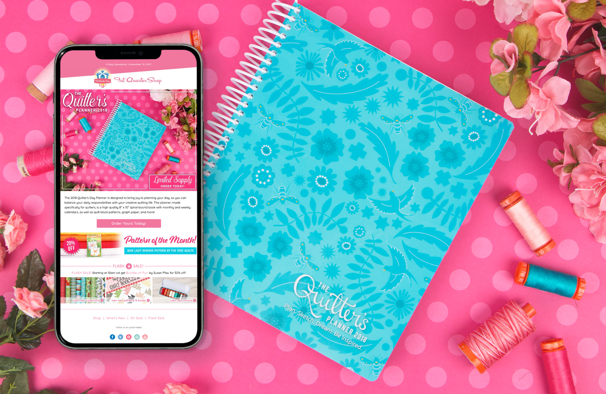 A blue coil-bound planner with a floral pattern on the cover. The book is sitting on a pink polka-dotted background and is surrounded by vibrant spools of thread and pink flowers. A phone is superimposed on top of the image, displaying an email that features the image behind to market this 2018 Quilters Planner. 