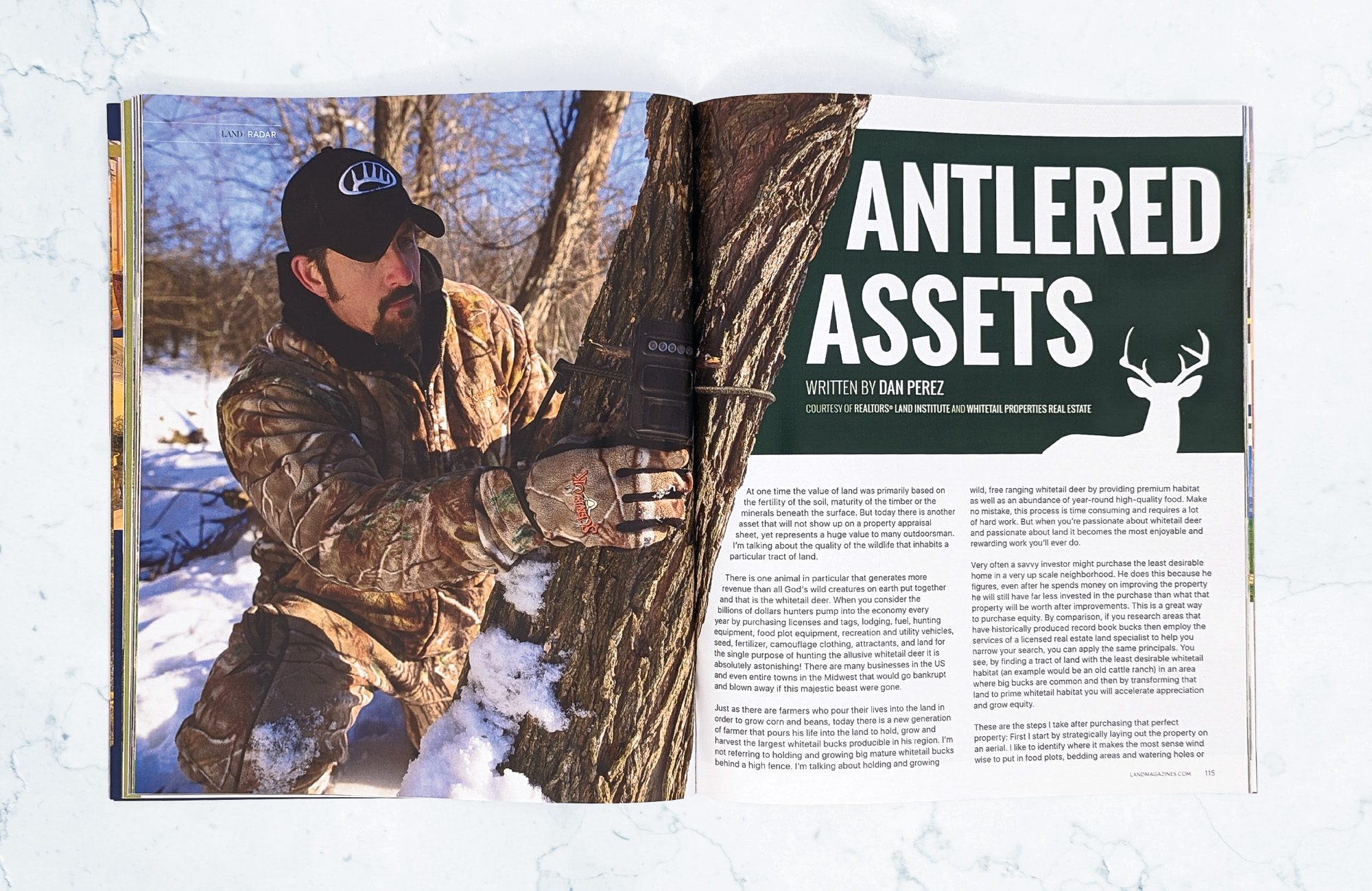 A magazine on a light marbled surface open to an editorial titled Antlered Assets. on the left is a picture of a bearded man in camo attire who is setting up a survey camera on a tree while kneeling in the snow. The tree juts out diagonally into the right page where the text for the editorial starts.