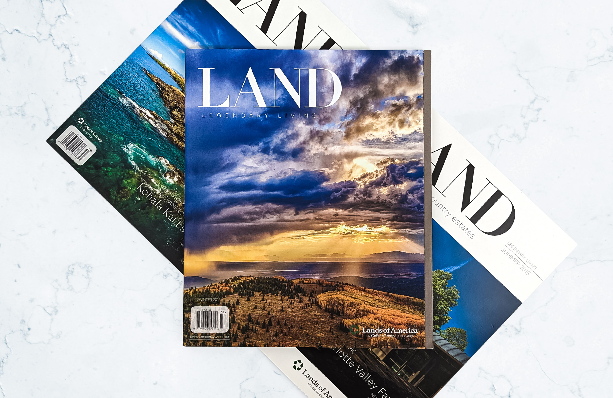 Three issues of LAND Magazine stacked on a marbled surface. The two magazines on the bottom are arranged diagonally and the one on top is parallel to the edge of the photo. The top magazine's cover depicts sunlight breaking through a cloudy sky and behind a mountain covered in trees. class=