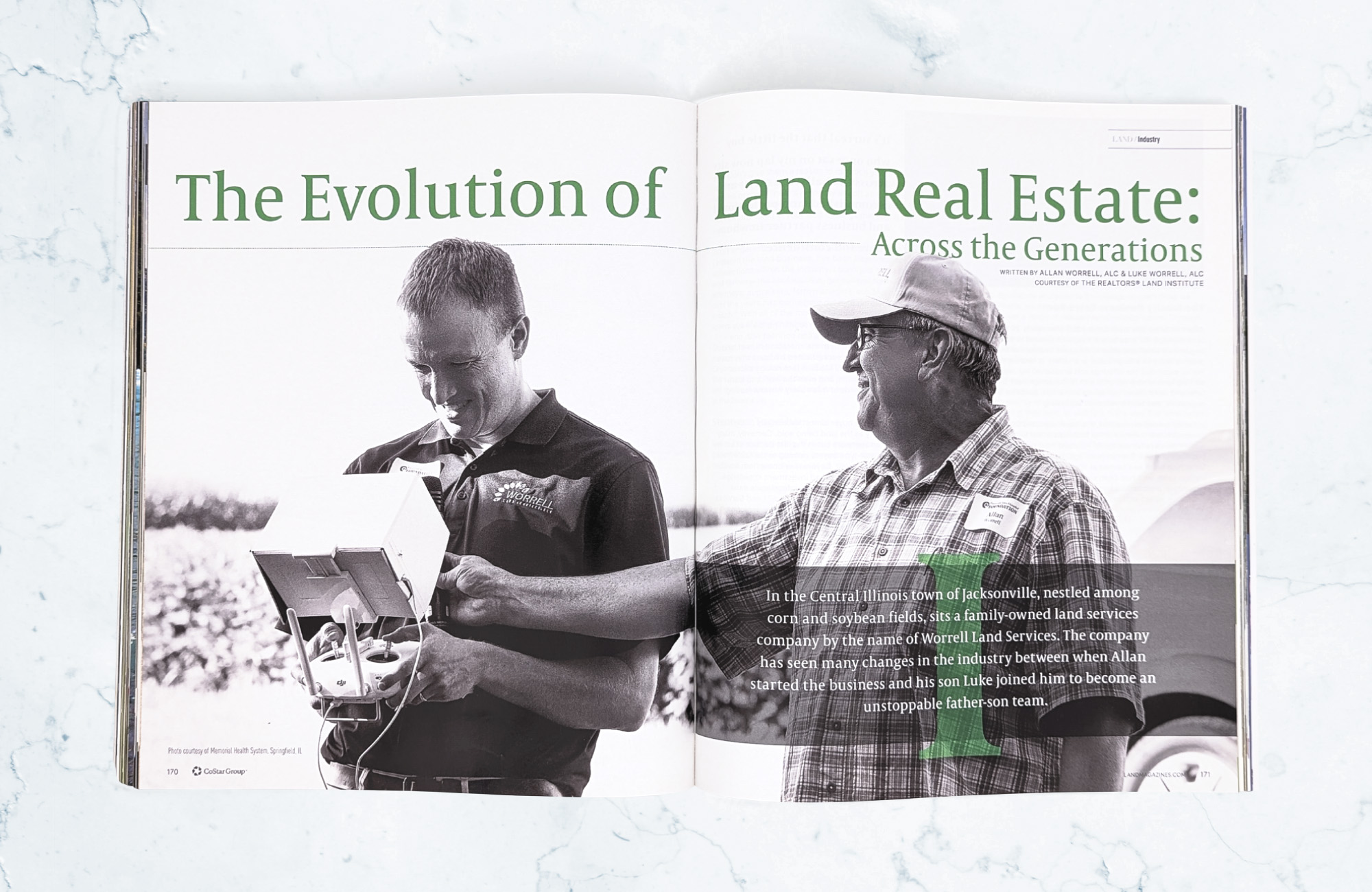 A magazine on a light marbled surface is open to a cover spread for an article titled The Evolution of Land Real Estate: Across the Generations. There is a black-and-white image of an older man in a baseball cap smiling as his adult son is holding a survey device.