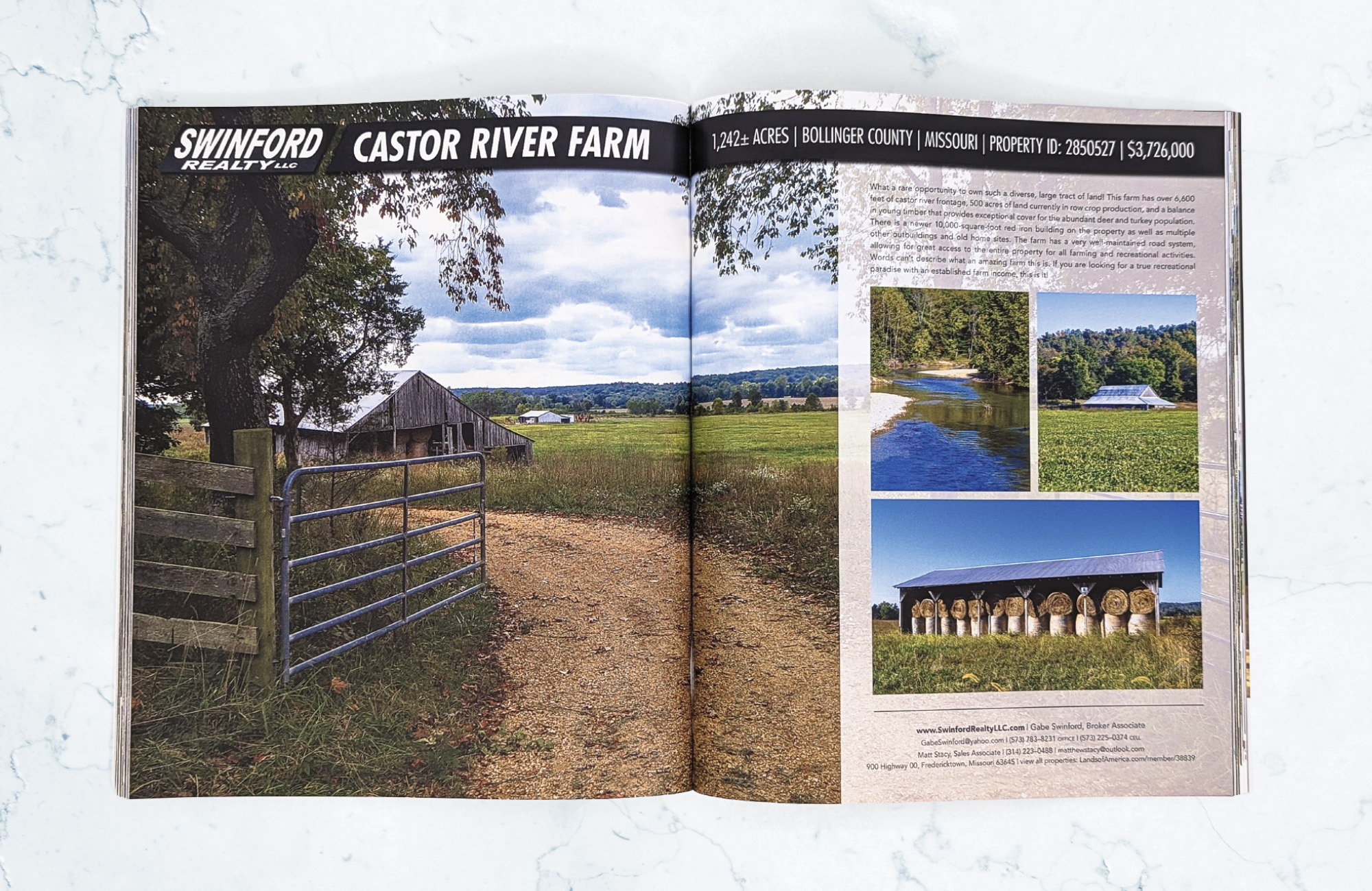 A magazine on a light marbled surface. The magazine is open to a showcase property listing, Castor River Farm, for Swinford Realty. The main photo spans across two pages and depicts an open metal gate and a dirt path leading to a barn. There is a description of the property on the right with a picture of a lake, a field, and a storage shed for hay bales.