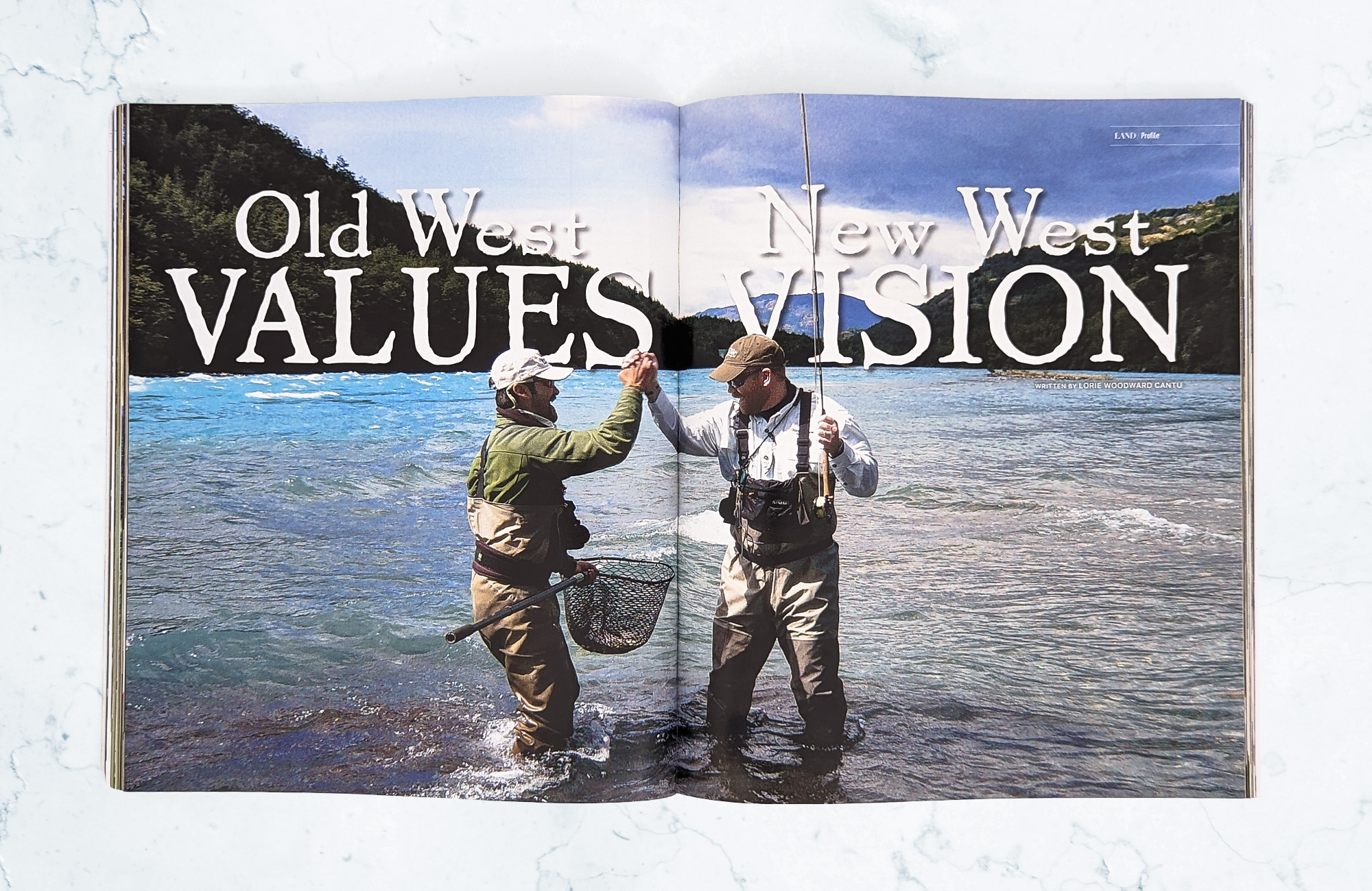 A magazine on a light marbled surface open to a cover spread for an editorial titled Old West Values, New West Vision. A photo takes up the whole spread with two men in knee-deep water clasping hands in the middle. The man on the left is holding a net while the man on the right is holding a fishing pole.