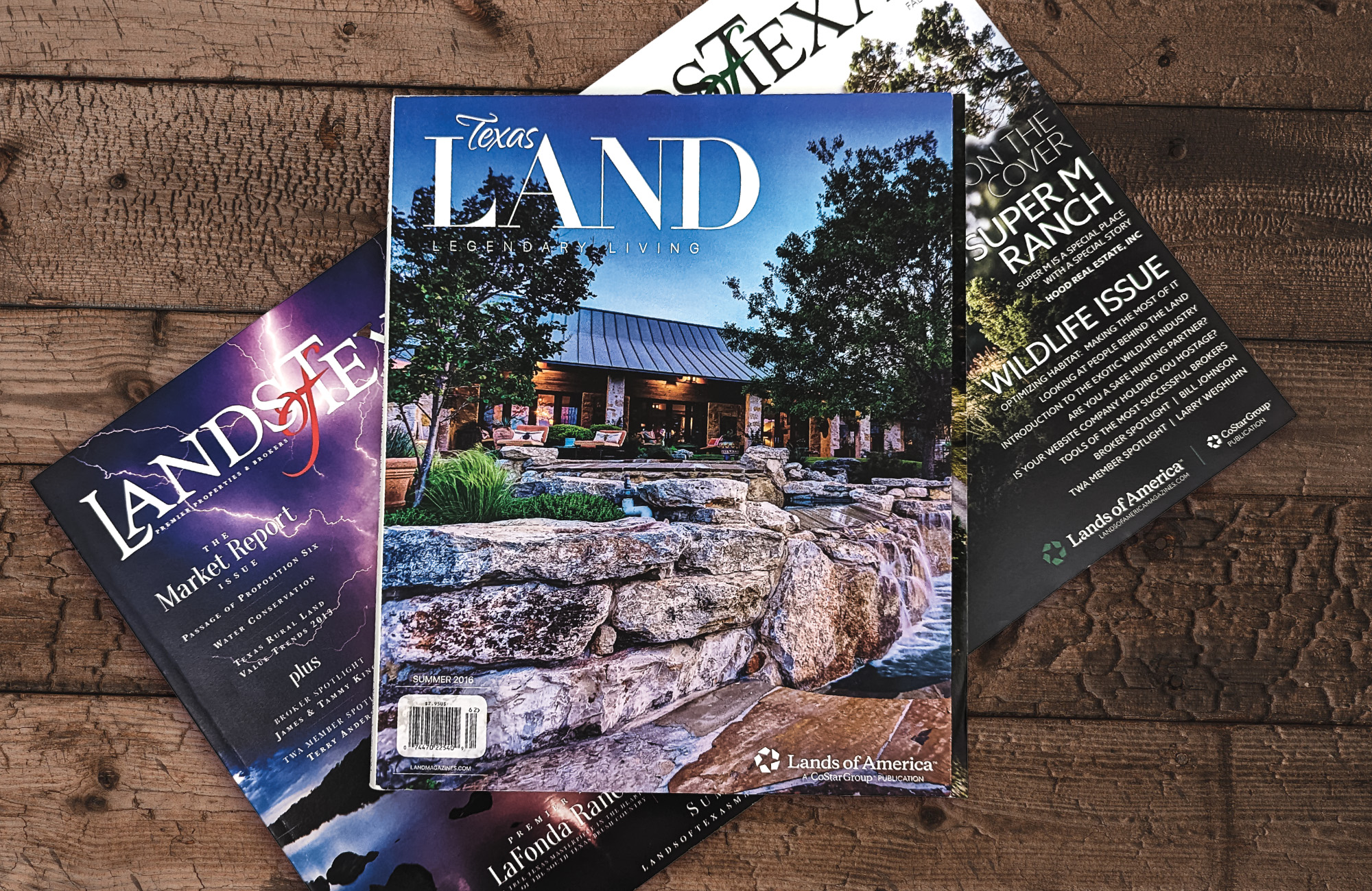One issue of Texas LAND Magazine stacked on top of two issues of Lands of Texas Magazine atop a rustic wooden surface. The cover of the top magazine is an image of a stone feature feeding into a swimming pool with a luxurious furnished patio in the background.  class=