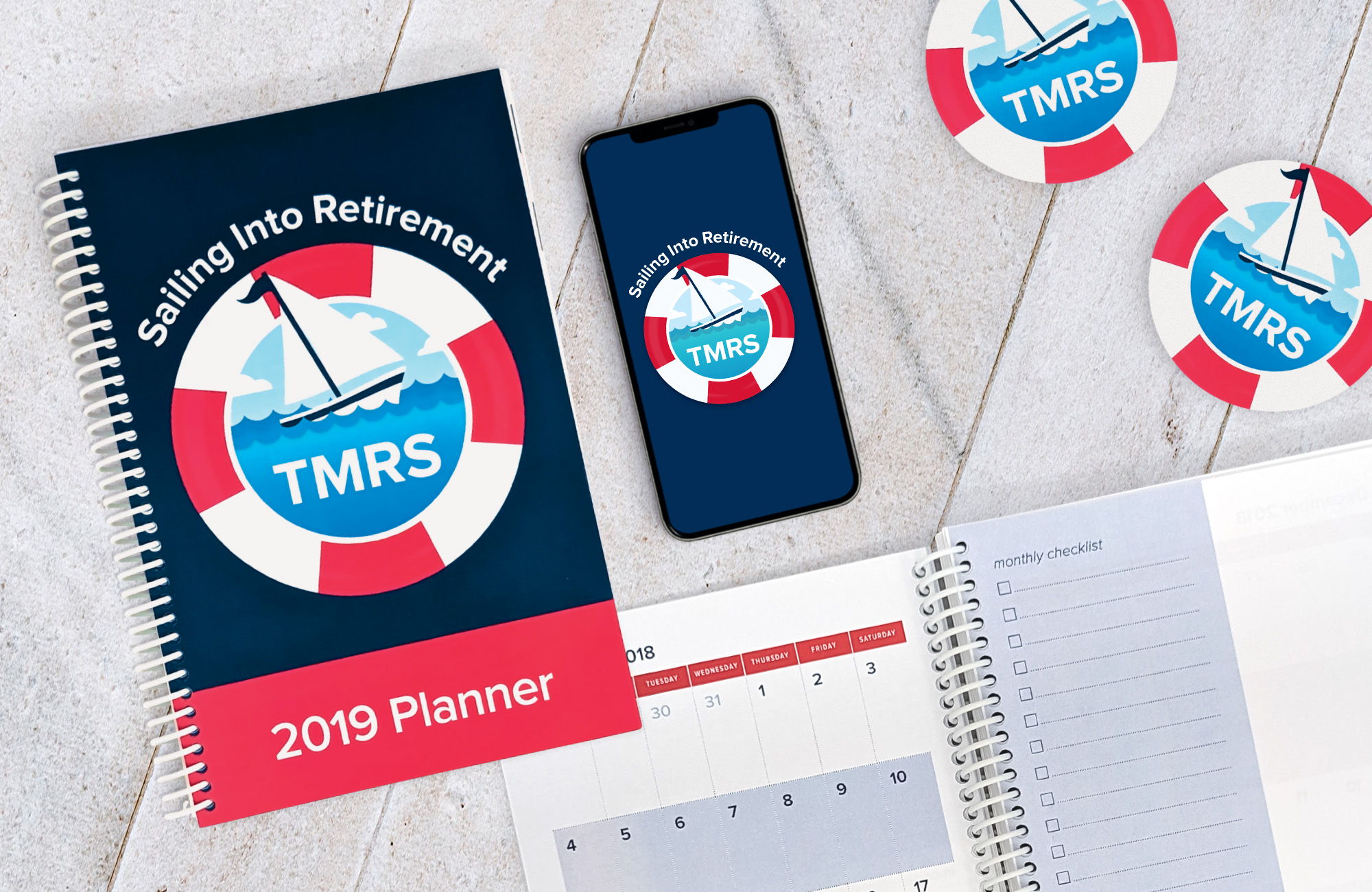 A 2019 coil-bound planner with a graphic resembling a sailboat encircled by a life preserver on the cover. Above the graphic reads the phrase Sailing Into Retirement. A phone open to the splash screen of an app and two coasters featuring the same graphic are visible to the right of the planner. A second planner is open to a monthly calendar of November 2018, but is partially obscured by the other planner. class=