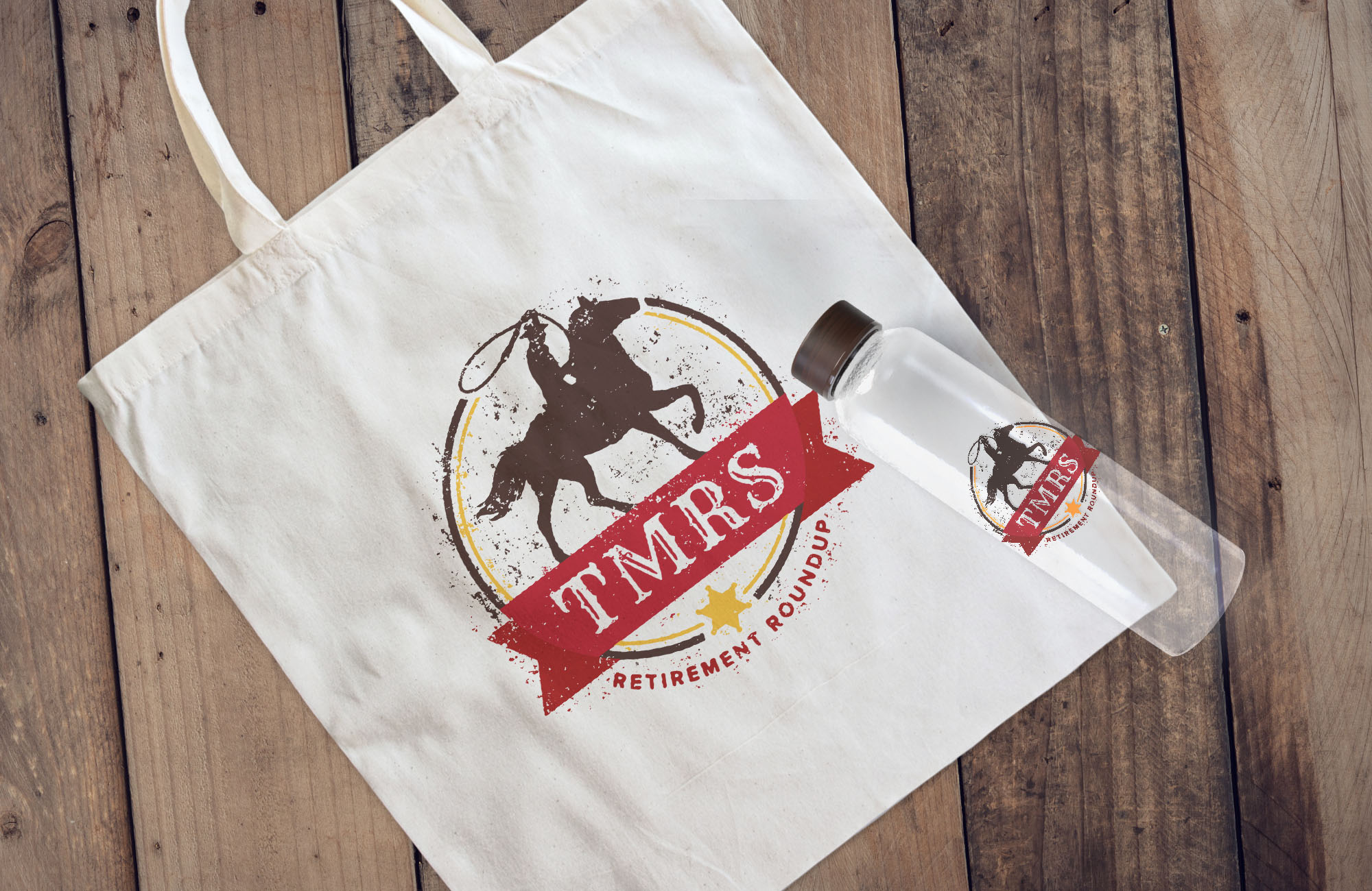 A tote bag and a glass water bottle sit on a wooden surface. Both items feature the Retirement Roundup emblem, a cowboy on a horse above a ribbon that reads TMRS.